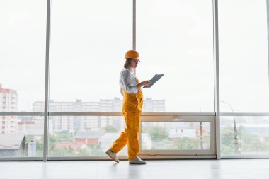 Büyük pencerenin yanında. Sarı üniformalı kadın işçi ya da mühendis ve içeride duran sert şapkalı..