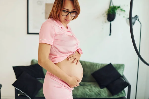 穿着粉色睡衣和眼镜站在室内的年轻孕妇白天 — 图库照片