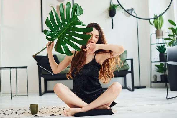 Young woman in pajamas sitting on the floor indoors at daytime and holding big green leaf.