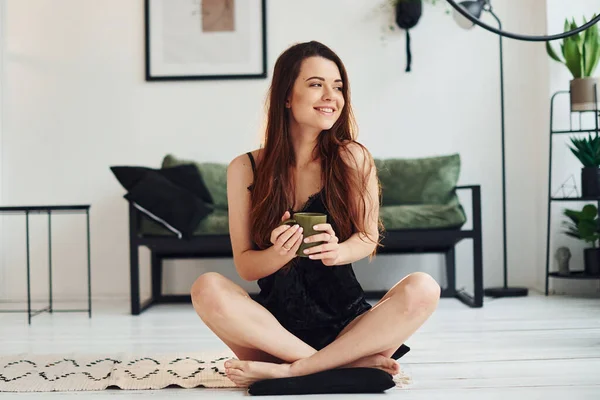 Young woman in pajamas sitting on the floor indoors at daytime with cup of drink.