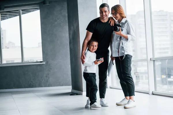 Resmi giyinmiş bir kadın ve elinde kamerayla boş bir odada bir adam ve küçük bir çocukla duruyor..