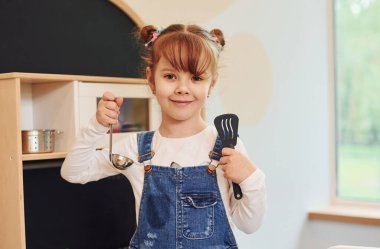Aletleri elinde tutuyor ve gösteriyor. Günlük kıyafetli küçük kız mutfakta oyuncaklarla oynayarak eğleniyor..
