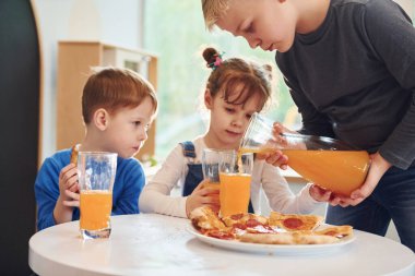 Üç çocuk masada oturuyor ve birlikte portakal suyu ile pizza yiyorlar..