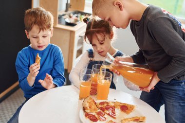 Üç çocuk masada oturuyor ve birlikte portakal suyu ile pizza yiyorlar..