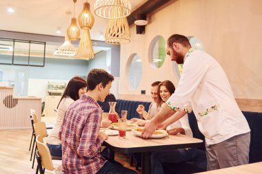 Şef yemek getiriyor. Evde oturup pizza yiyen bir grup genç arkadaş..