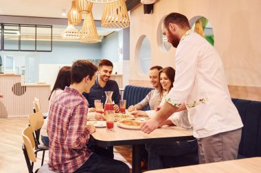 Şef yemek getiriyor. Evde oturup pizza yiyen bir grup genç arkadaş..