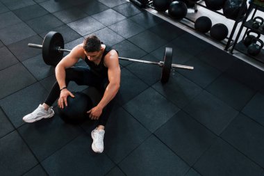 Spor elbiseli güçlü bir adam spor salonunda yerde ekipmanlarıyla oturuyor..
