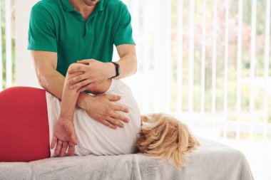 Yaşlı kadın kliniği ziyaret ediyor, uzanıyor ve rehabilitasyon görüyor. Erkek eğitmen ona yardım ediyor..