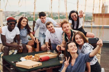 Kız selfie çekiyor. Lezzetli pizzayla. Gündelik giysiler içinde bir grup genç gündüz vakti çatıda parti veriyor..