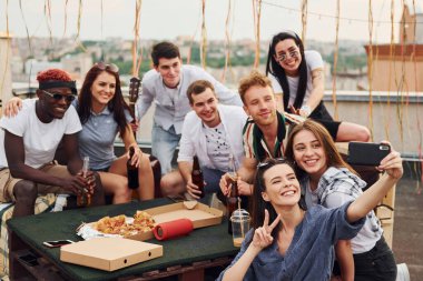 Kız selfie çekiyor. Lezzetli pizzayla. Gündelik giysiler içinde bir grup genç gündüz vakti çatıda parti veriyor..