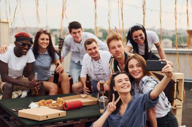 Kız selfie çekiyor. Lezzetli pizzayla. Gündelik giysiler içinde bir grup genç gündüz vakti çatıda parti veriyor..