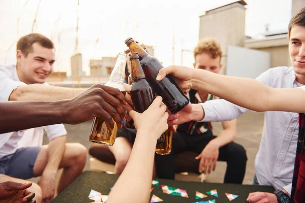 Doing cheers by bottles with beer. Group of young people in casual clothes have a party at rooftop together at daytime.