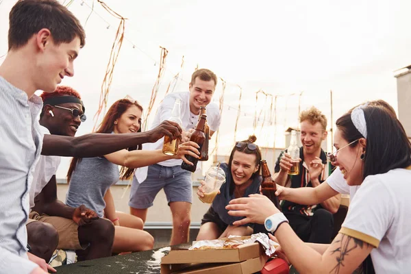 Doing cheers by bottles with beer. Group of young people in casual clothes have a party at rooftop together at daytime.