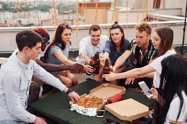 Doing cheers by bottles with beer. Group of young people in casual clothes have a party at rooftop together at daytime.