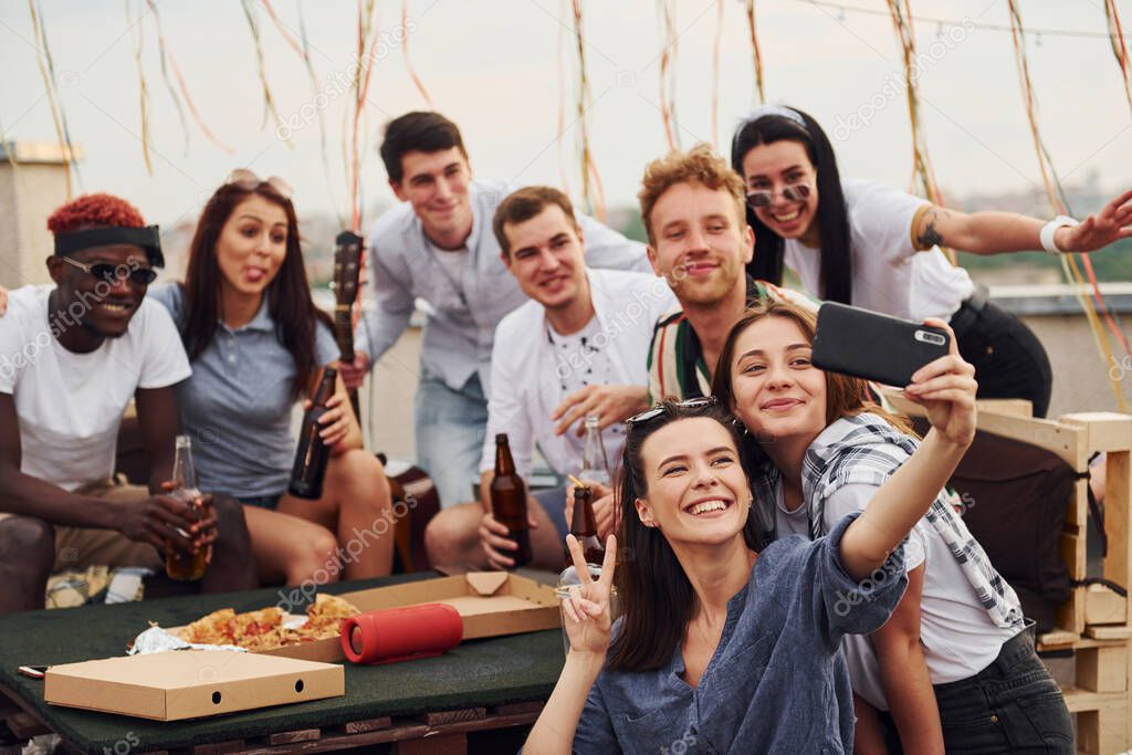 Girl making selfie. With delicious pizza. Group of young people in casual clothes have a party at rooftop together at daytime.
