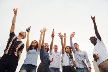 Yüksekte duruyor ve bulutlu gökyüzüne bakıyor. Gündelik giysiler içinde bir grup genç gündüz vakti çatıda parti veriyor..