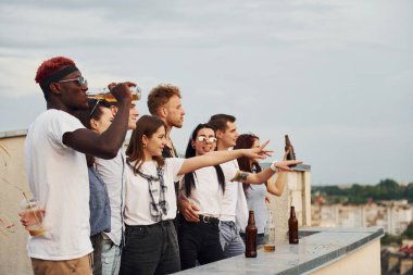 Alkolle yüksekte duruyor. Bulutlu gökyüzü. Gündelik giysiler içinde bir grup genç gündüz vakti çatıda parti veriyor..