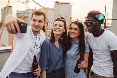 Gündelik giysiler içindeki bir grup genç gündüzleri çatıda parti veriyor ve telefonla selfie çekiyorlar..