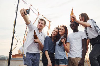 Gündelik giysiler içindeki bir grup genç gündüzleri çatıda parti veriyor ve telefonla selfie çekiyorlar..