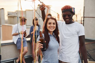 Kamera için poz vermek. Gündelik giysiler içinde bir grup genç gündüz vakti çatıda parti veriyor..