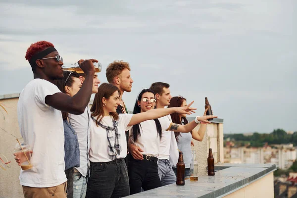 Alkolle yüksekte duruyor. Bulutlu gökyüzü. Gündelik giysiler içinde bir grup genç gündüz vakti çatıda parti veriyor..
