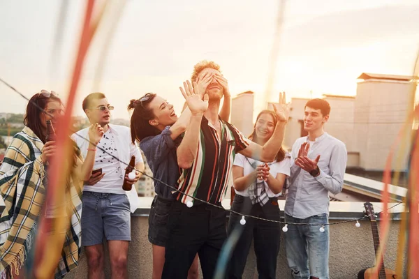 Playing game. Man\'s eyes covered by hands. Group of young people in casual clothes have a party at rooftop together at daytime.