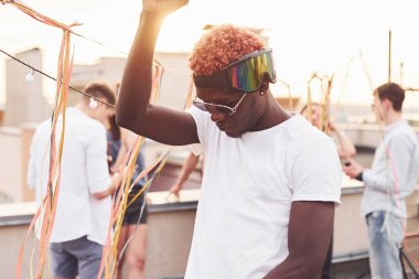 Gülümseyen ve dans eden Afrikalı Amerikalı bir adam. Gündelik giysiler içinde bir grup genç gündüz vakti çatıda parti veriyor..