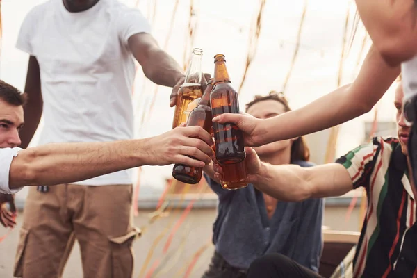 Doing cheers by bottles of beer. Group of young people in casual clothes have a party at rooftop together at daytime.