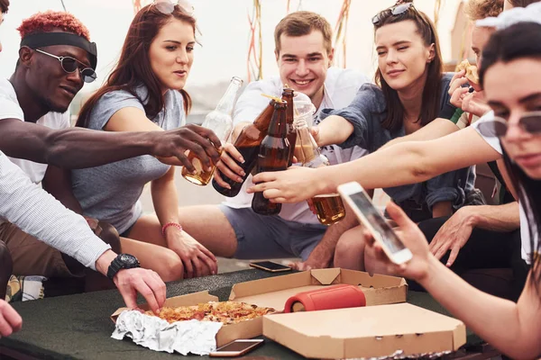 Lezzetli pizza. Gündelik giysiler içinde bir grup genç gündüz vakti çatıda parti veriyor..