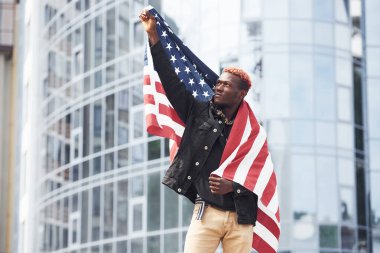 ABD bayrağı vatanseverlerin elinde. Gurur ve özgürlük kavramı. Şehirdeki siyah ceketli genç Afro-Amerikan adam modern iş binasına karşı duruyor..