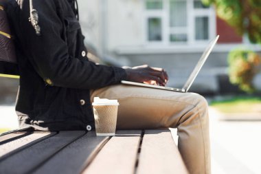 Dizüstü bilgisayarla çalışıyor. Siyah ceketli Afro-Amerikan 'ın yakın görüntüsü. Kentin dışında bankta oturuyor..