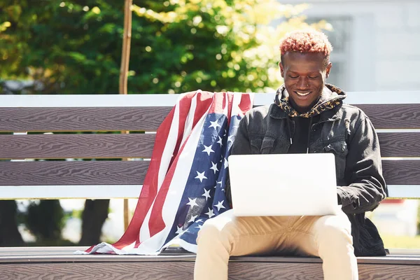 USA flag leaning behind. Using laptop. Young african american man in black jacket outdoors in the city sitting on bench.