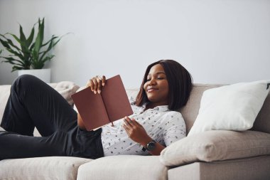 Ellerinde not defteriyle kanepeye uzanmış beyaz gömlekli Afro-Amerikan kadın..