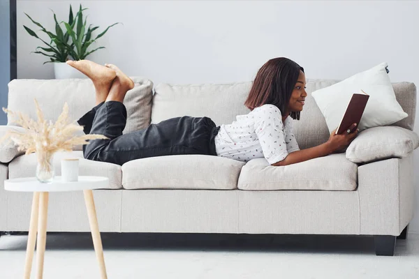 Positive african american woman in white shirt lying down on sofa with notepad in hands.