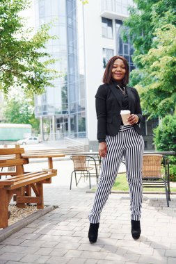 Kahve molası. Şehirde, yeşil ağaçların yanında ve iş inşaatına karşı şık giyinmiş Afro-Amerikalı genç bir kadın..