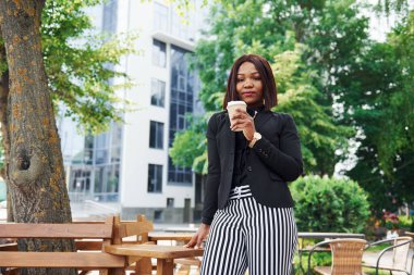 Kahve molası. Şehirde, yeşil ağaçların yanında ve iş inşaatına karşı şık giyinmiş Afro-Amerikalı genç bir kadın..