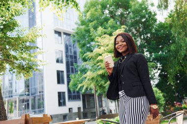 Kahve molası. Şehirde, yeşil ağaçların yanında ve iş inşaatına karşı şık giyinmiş Afro-Amerikalı genç bir kadın..