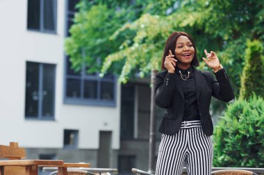 Telefonla konuşuyorum. Şehirde, yeşil ağaçların yanında ve iş inşaatına karşı şık giyinmiş Afro-Amerikalı genç bir kadın..