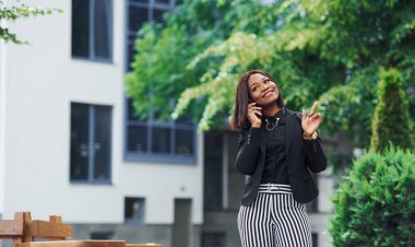 Telefonla konuşuyorum. Şehirde, yeşil ağaçların yanında ve iş inşaatına karşı şık giyinmiş Afro-Amerikalı genç bir kadın..