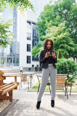 Telefonu bekletiyorum. Şehirde, yeşil ağaçların yanında ve iş inşaatına karşı şık giyinmiş Afro-Amerikalı genç bir kadın..
