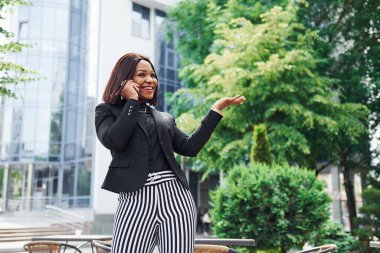 Telefonla konuşuyorum. Şehirde, yeşil ağaçların yanında ve iş inşaatına karşı şık giyinmiş Afro-Amerikalı genç bir kadın..