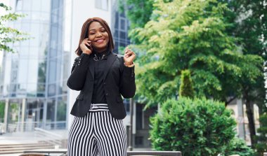 Telefonla konuşuyorum. Şehirde, yeşil ağaçların yanında ve iş inşaatına karşı şık giyinmiş Afro-Amerikalı genç bir kadın..