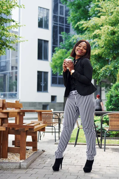 Kahve molası. Şehirde, yeşil ağaçların yanında ve iş inşaatına karşı şık giyinmiş Afro-Amerikalı genç bir kadın..