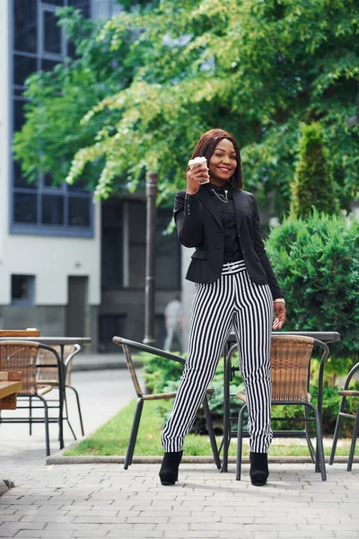 Kahve molası. Şehirde, yeşil ağaçların yanında ve iş inşaatına karşı şık giyinmiş Afro-Amerikalı genç bir kadın..