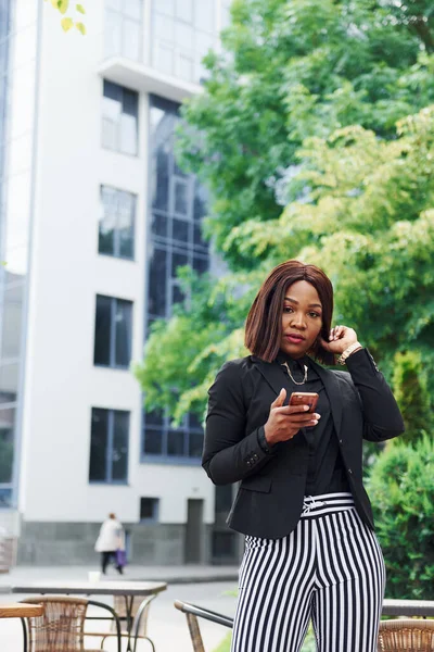 Telefonu bekletiyorum. Şehirde, yeşil ağaçların yanında ve iş inşaatına karşı şık giyinmiş Afro-Amerikalı genç bir kadın..