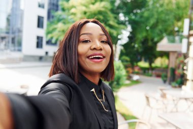 Selfie çekiyorum. Şehirde, yeşil ağaçların yanında ve iş inşaatına karşı şık giyinmiş Afro-Amerikalı genç bir kadın..