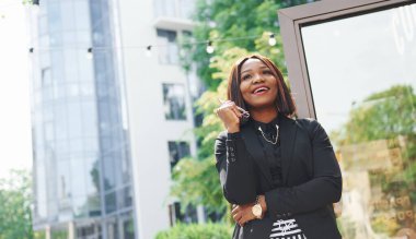 Bardakları elinde tutuyor. Şehirde, yeşil ağaçların yanında ve iş inşaatına karşı şık giyinmiş Afro-Amerikalı genç bir kadın..