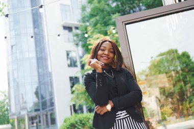 Bardakları elinde tutuyor. Şehirde, yeşil ağaçların yanında ve iş inşaatına karşı şık giyinmiş Afro-Amerikalı genç bir kadın..