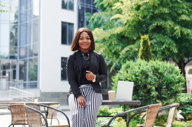 Laptop ve masa. Şehirde, yeşil ağaçların yanında ve iş inşaatına karşı şık giyinmiş Afro-Amerikalı genç bir kadın..