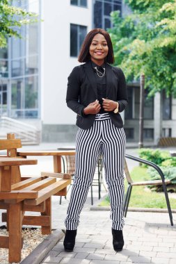 Şehirde, yeşil ağaçların yanında ve iş inşaatına karşı şık giyinmiş Afro-Amerikalı genç bir kadın..